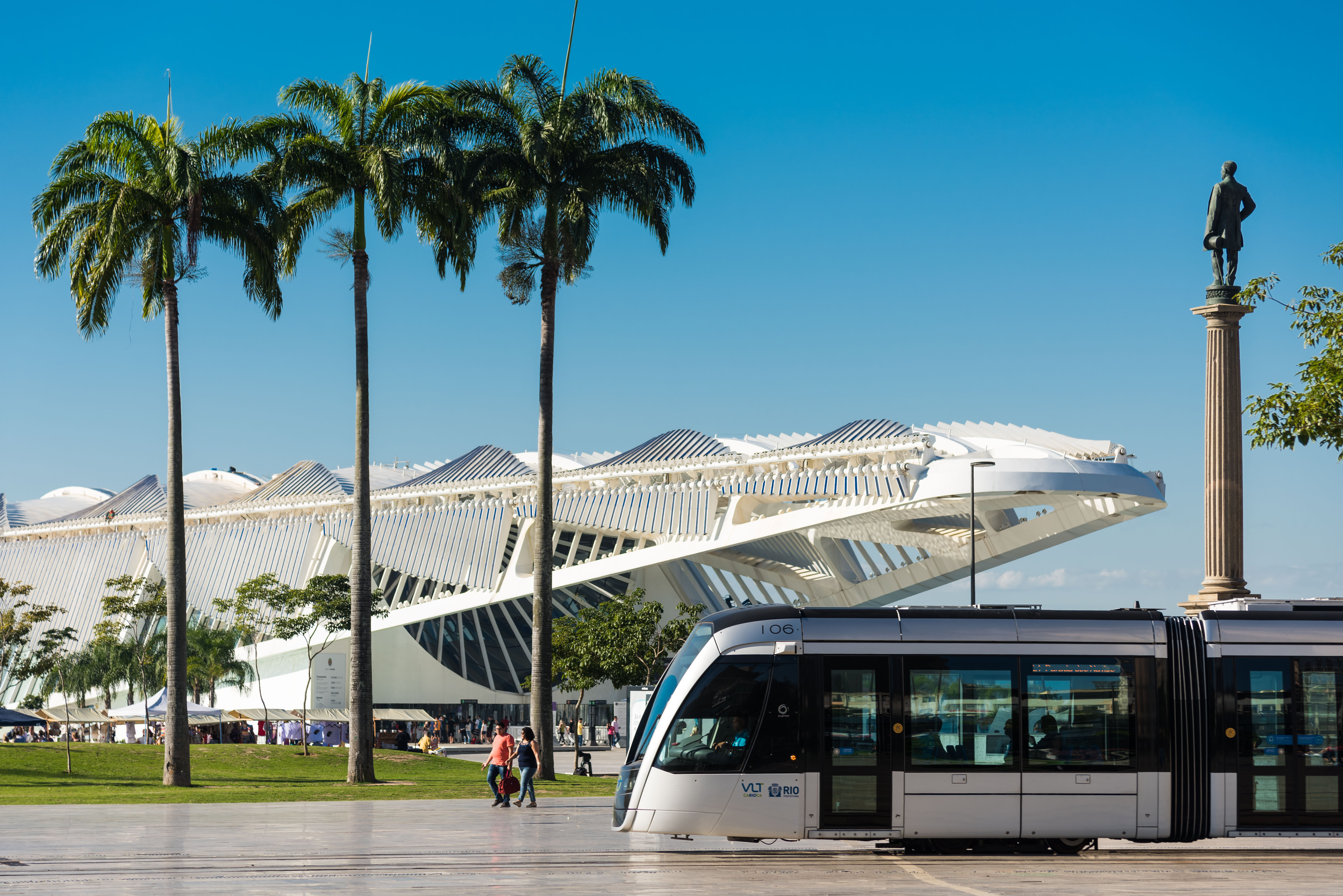 Veículo leve sobre trilhos circulando próximo ao Museu do Amanhã, com palmeiras e pedestres em um dia ensolarado.